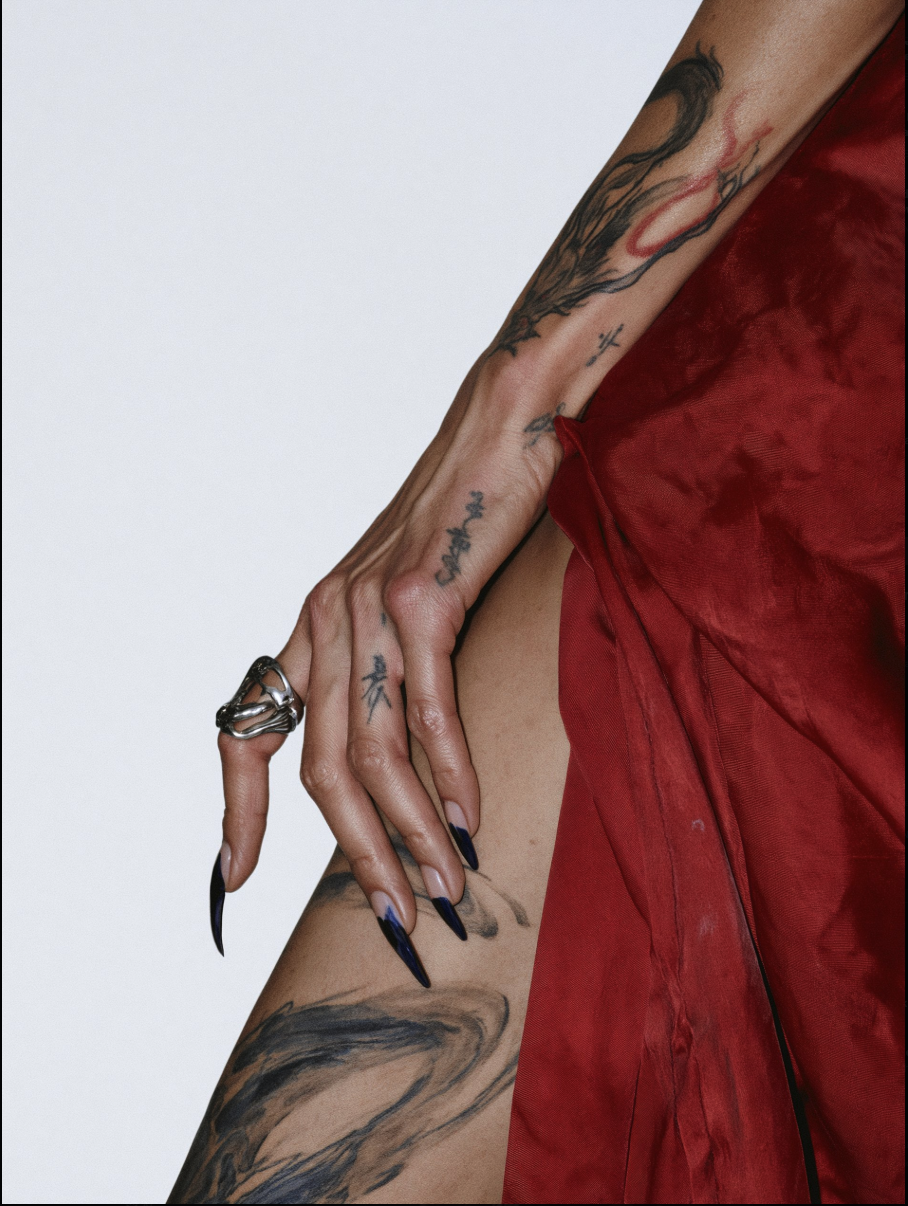 A close-up of a tattooed hand with long black pointed nails resting against draped red fabric.