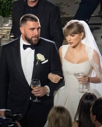 A woman resembling Taylor Swift in a white wedding dress walking beside a man resembling Travis Kelce in a black tuxedo at an indoor event.