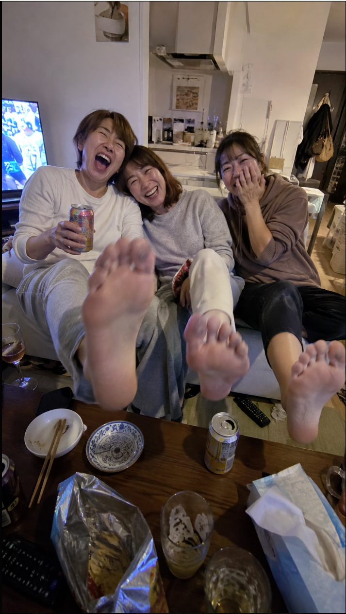 Three women laughing together on a couch in a home interior, with their bare feet extended toward the camera.