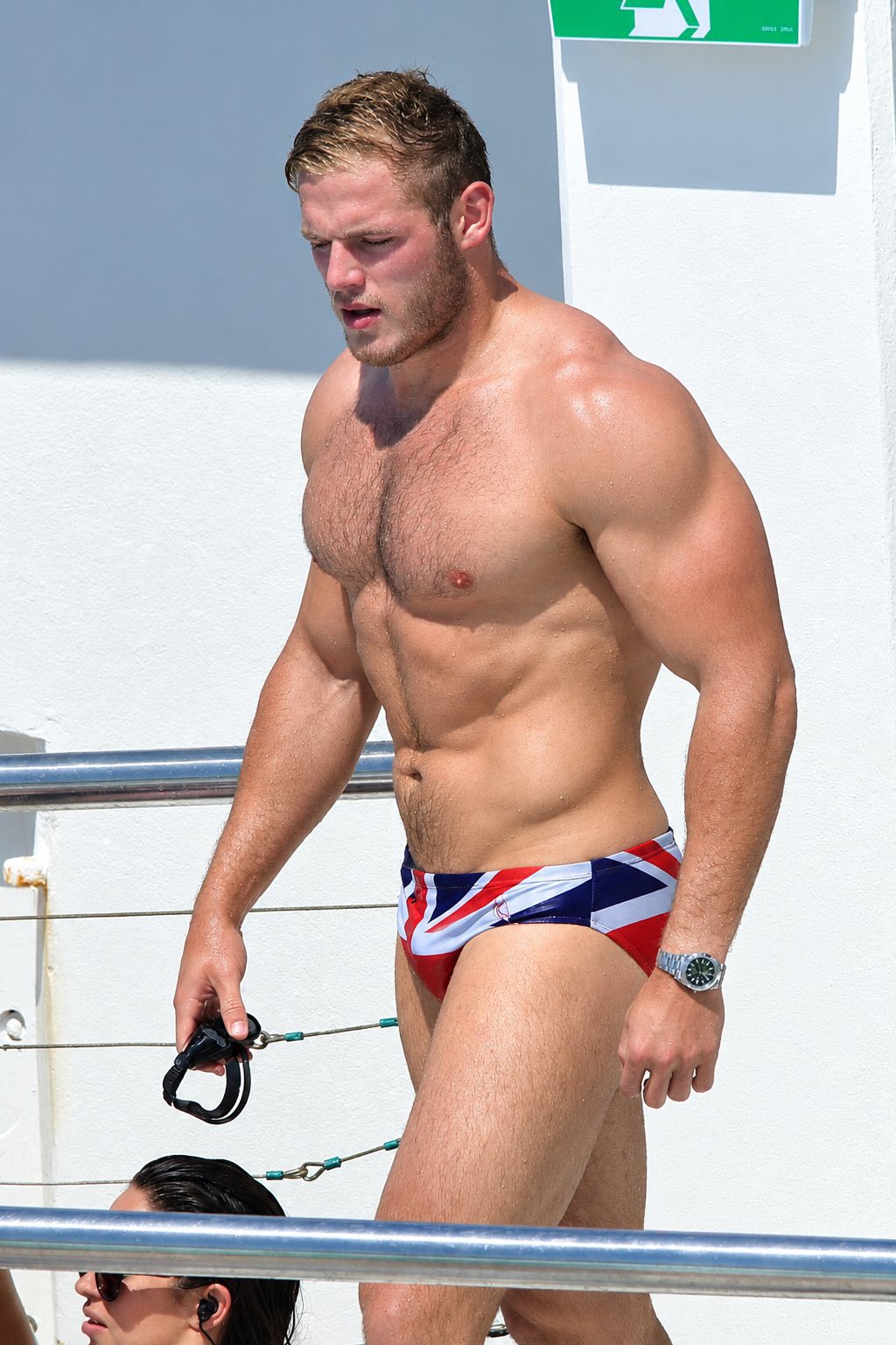A muscular man resembling Tom Burgess wearing Union Jack-patterned swim briefs on a boat.