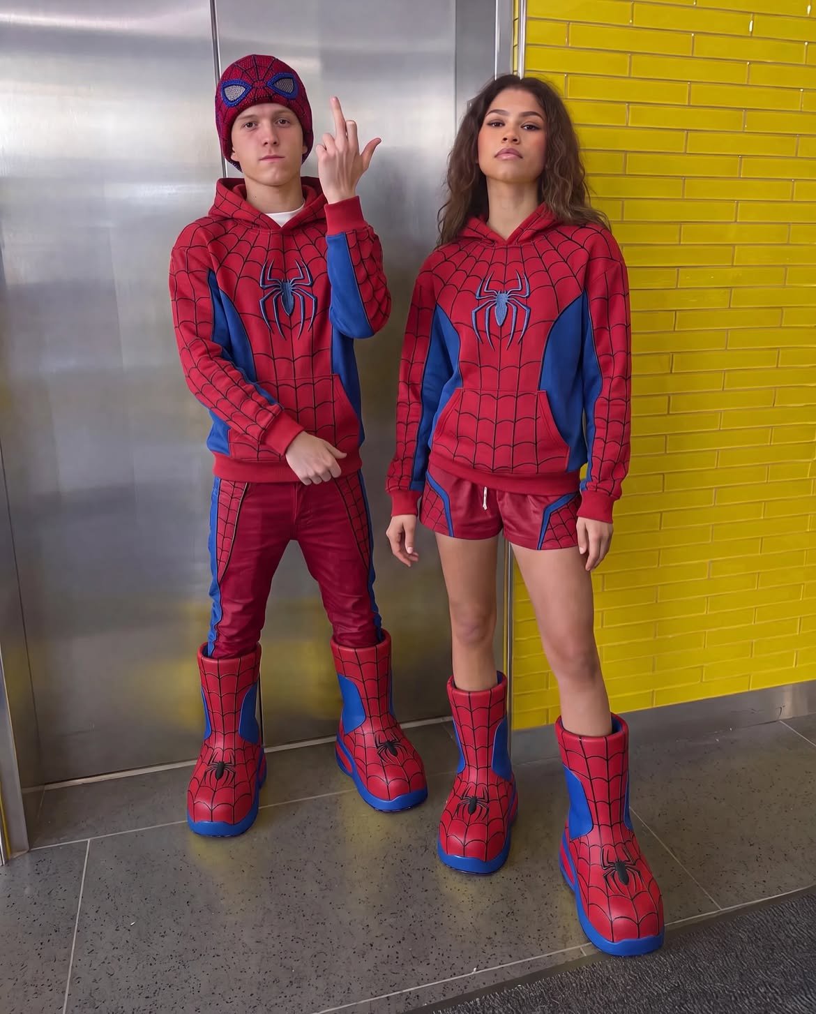 A man and a woman resembling Tom Holland and Zendaya standing side by side in matching red and blue Spider-Man hoodies, pants or shorts, and oversized boots.