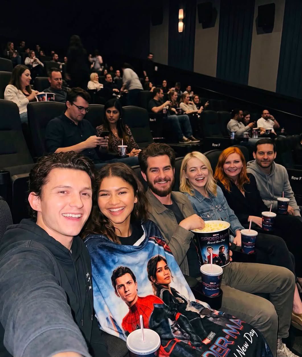People resembling Tom Holland, Zendaya, Andrew Garfield, and others seated together in a movie theater holding Spider-Man: Brand New Day popcorn buckets.