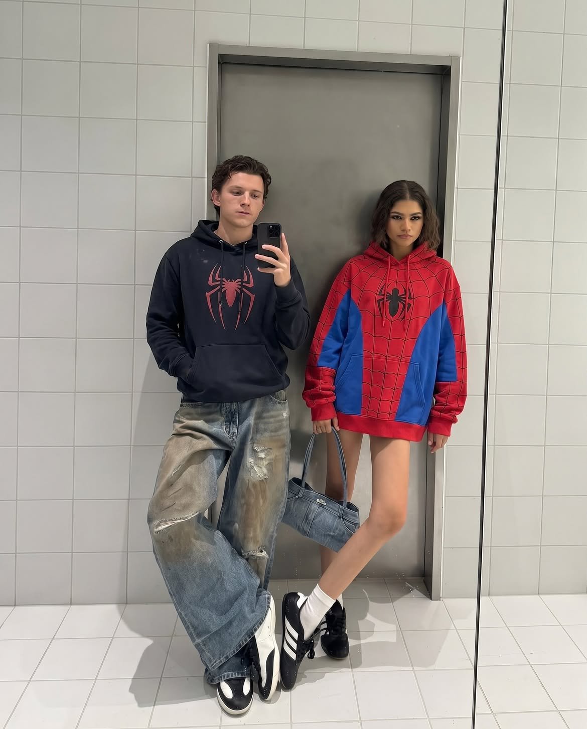 A man resembling Tom Holland and a woman resembling Zendaya in Spider-Man hoodies taking a mirror selfie.