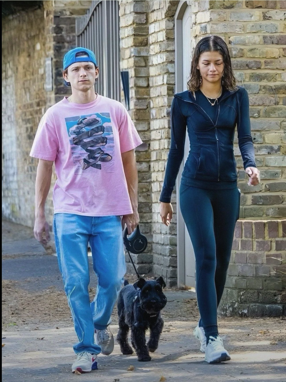 A man resembling Tom Holland and a woman resembling Zendaya walking a black dog along a brick-walled alley.