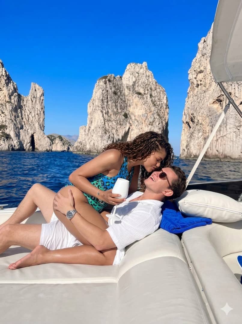 Two people resembling Tom Holland and Zendaya relaxing together on a yacht near rocky coastal cliffs.