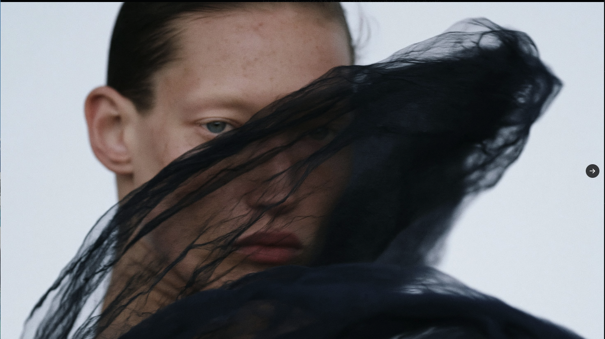 A close-up portrait , her face partially covered by a sheer black veil against a light background.