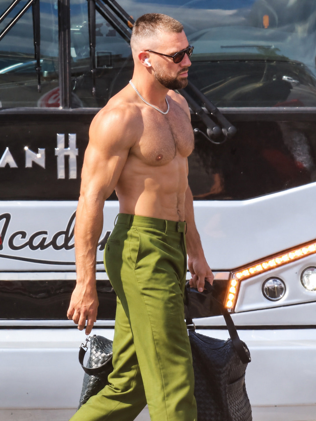 A shirtless, muscular man resembling Travis Kelce walking outdoors carrying a bag, with a tour bus behind him.