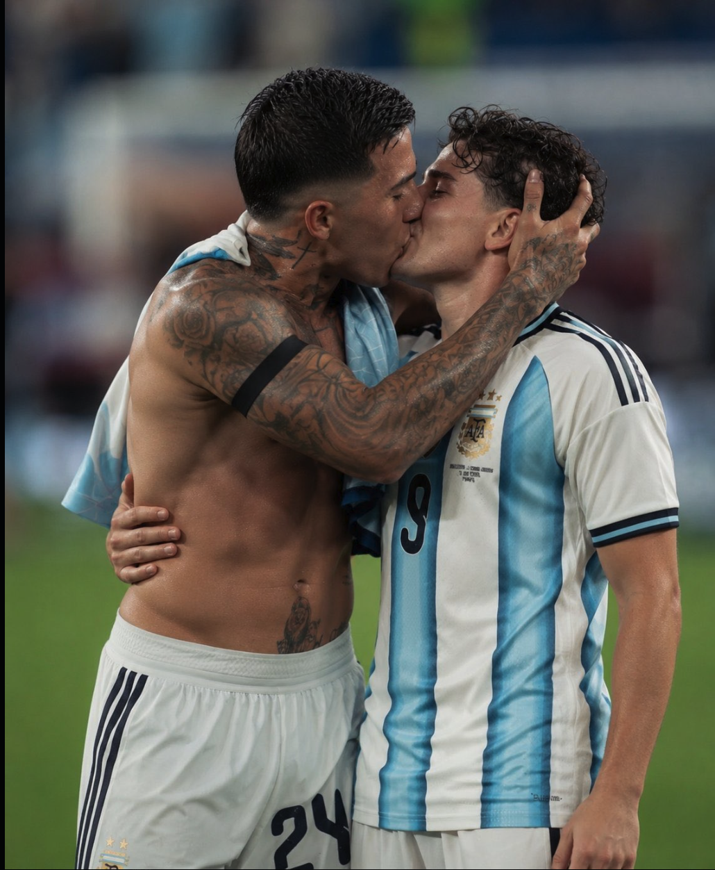 Two men on a soccer pitch kissing, one shirtless with tattoos and an Argentine flag draped over his shoulder, the other wearing an Argentina jersey numbered 9.