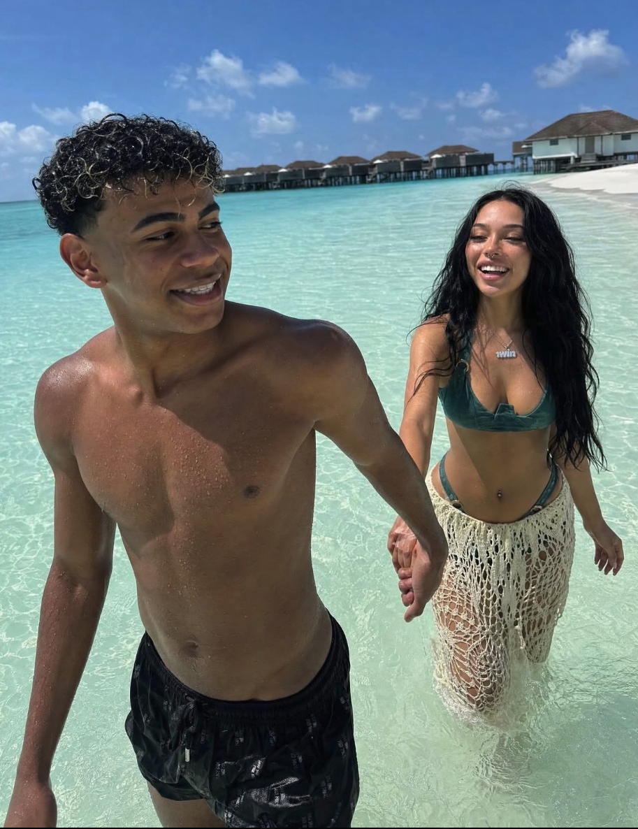 Lamine Yamal and a young woman in swimwear standing in shallow turquoise water with overwater bungalows in the background; the woman wears a '1win' pendant.