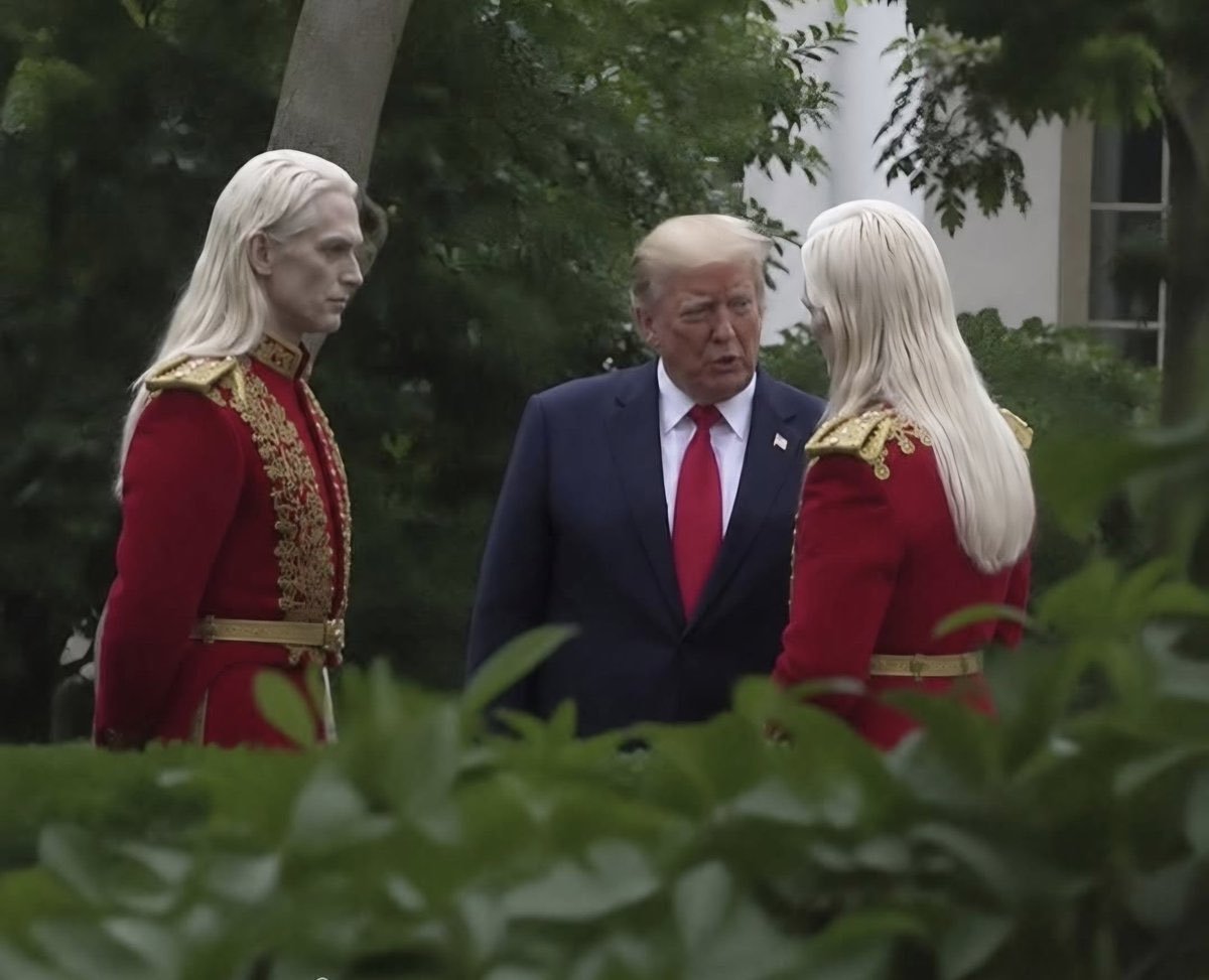 Donald Trump in a dark suit speaking with two pale-haired figures in red military-style uniforms amid garden foliage.