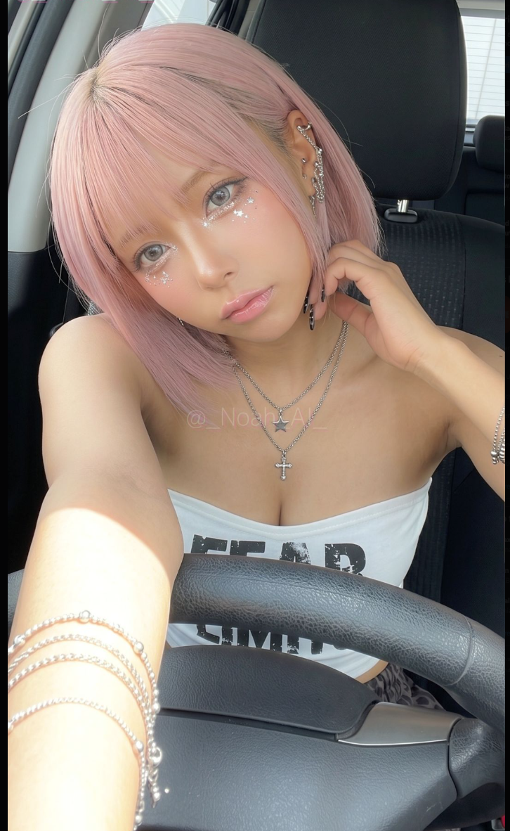 A woman taking a selfie in a car, with pink hair, glossy makeup, and a white tube top.