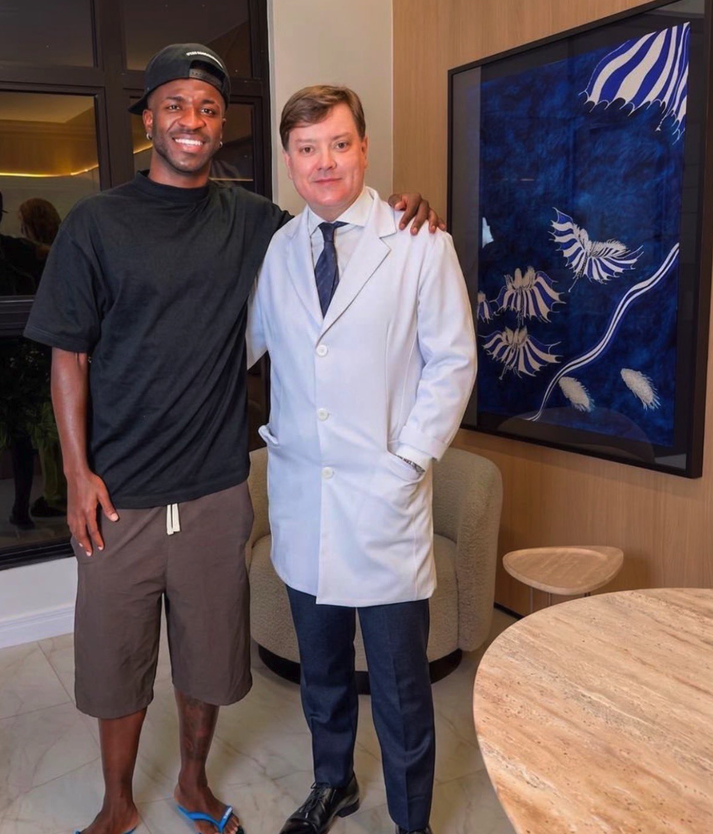 Two men posing together indoors, one in casual clothing and one wearing a white lab coat, with a large blue botanical artwork behind them.