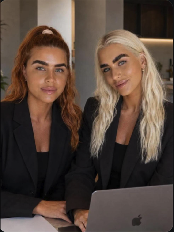 Two women with long hair wearing black blazers, seated indoors beside an open laptop.