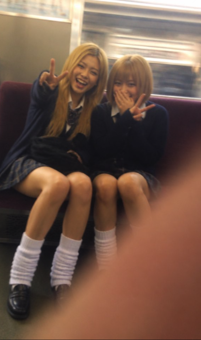 Two young women in dark school uniforms smiling and making peace signs while seated on a train.