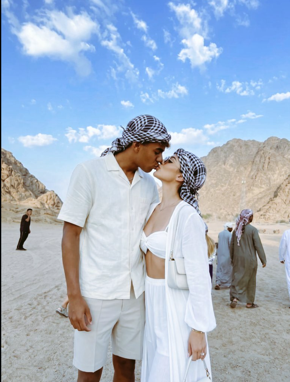 A woman and Lamine Yamal in white outfits and checkered headscarves kissing outdoors in a desert mountain landscape.