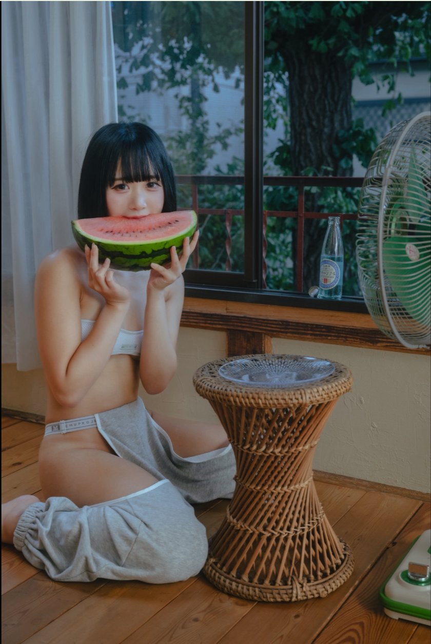 A young woman with short dark hair seated on a wooden floor, holding a slice of watermelon near her face.
