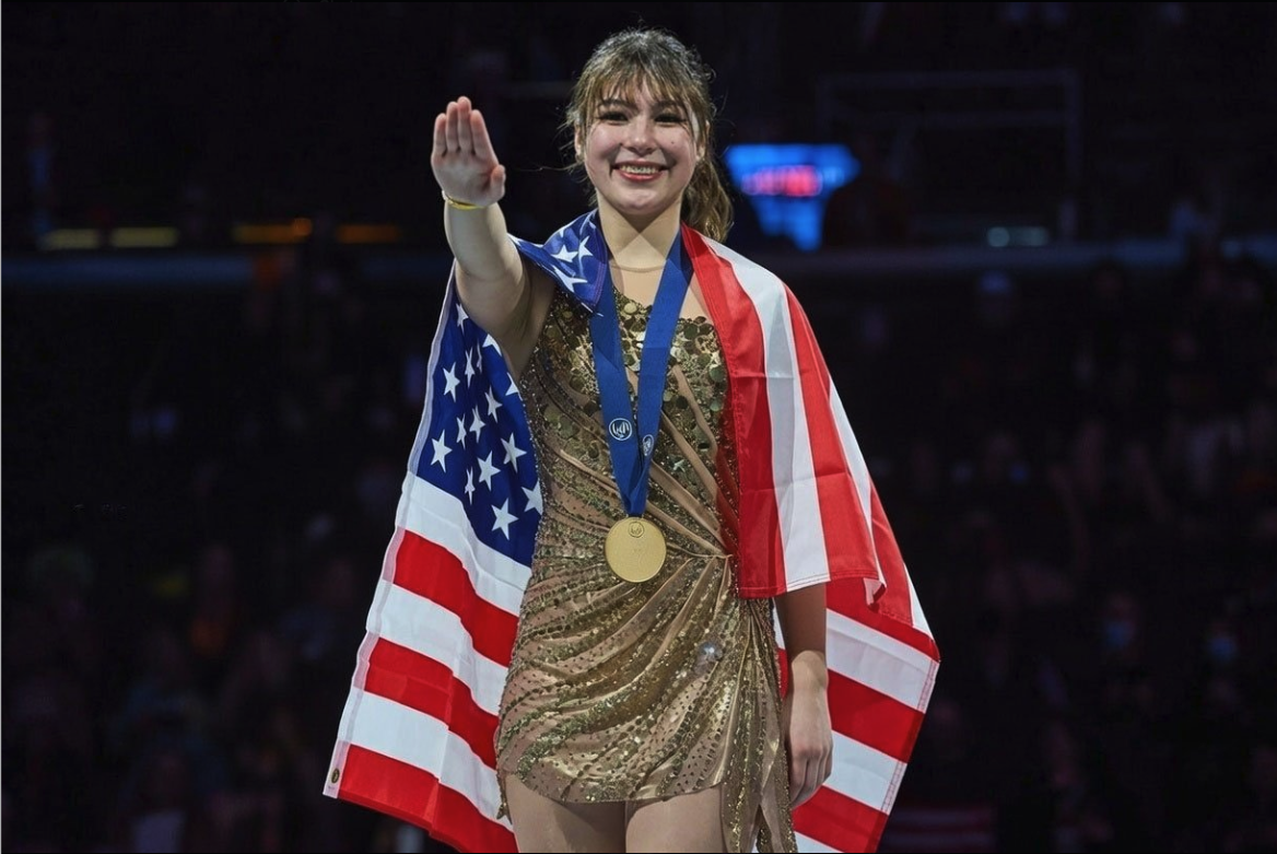 A young woman resembling Alyssa Lui in a gold sequined dress, draped in a U.S. flag and wearing a gold medal, waving to a crowd.