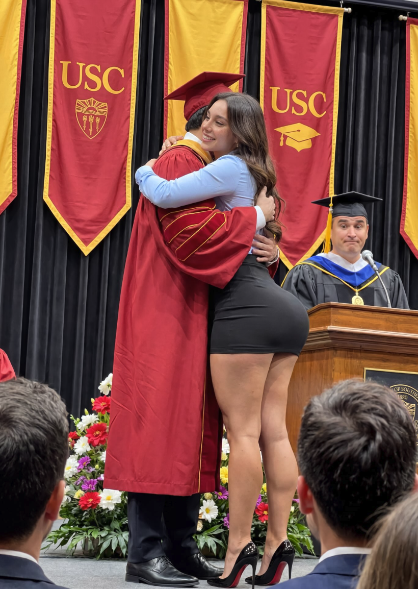 A woman in a blue blouse and short black skirt embracing a graduate in red cap and gown in front of USC banners.