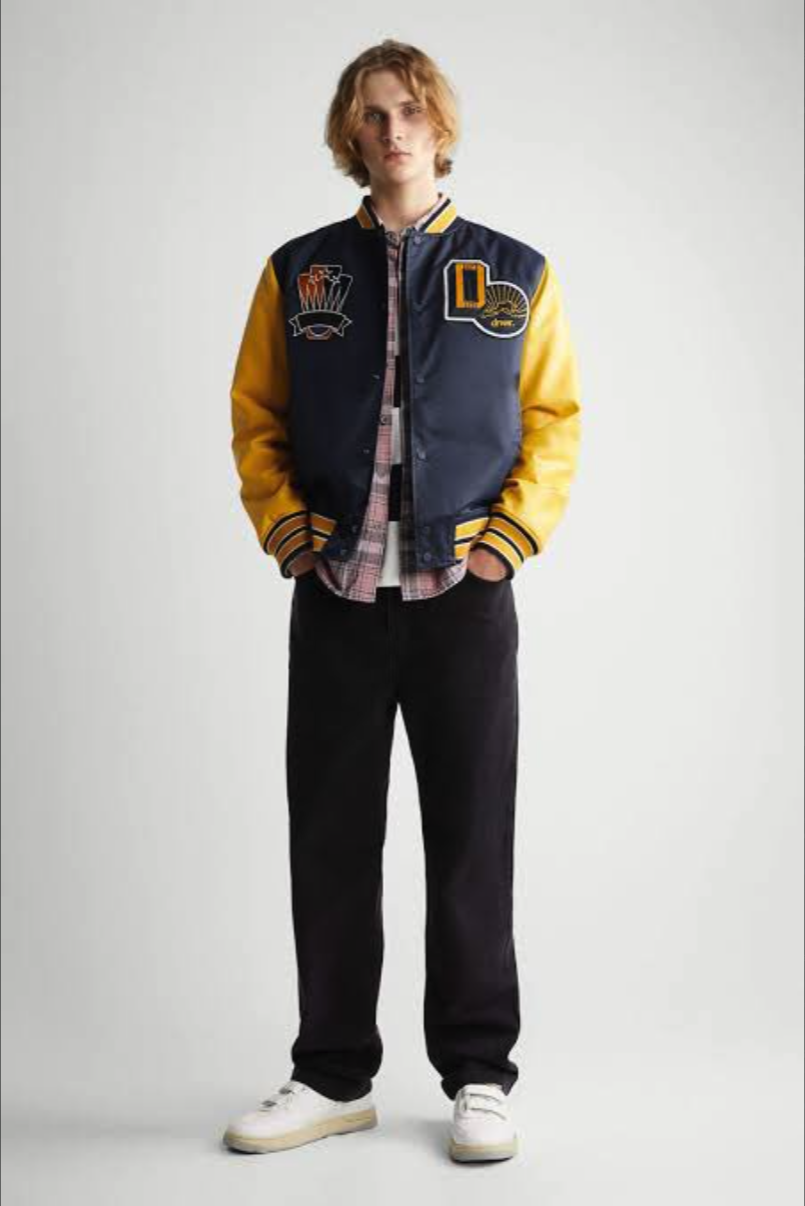 A man with shoulder-length hair wearing a navy and yellow varsity jacket with patches, a plaid shirt, dark pants, and white sneakers.