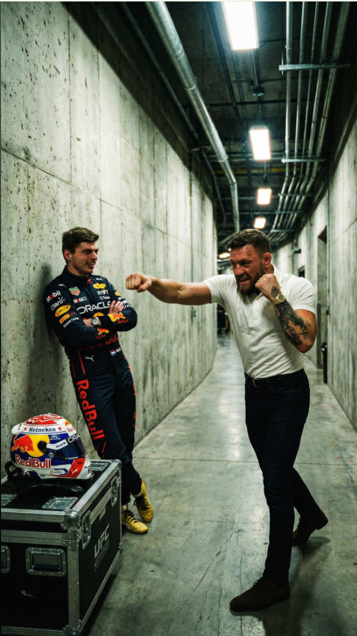 Two men resembling Max Verstappen and Conor McGregor in a service corridor, one in Red Bull Racing gear and the other in a white polo with fists raised.