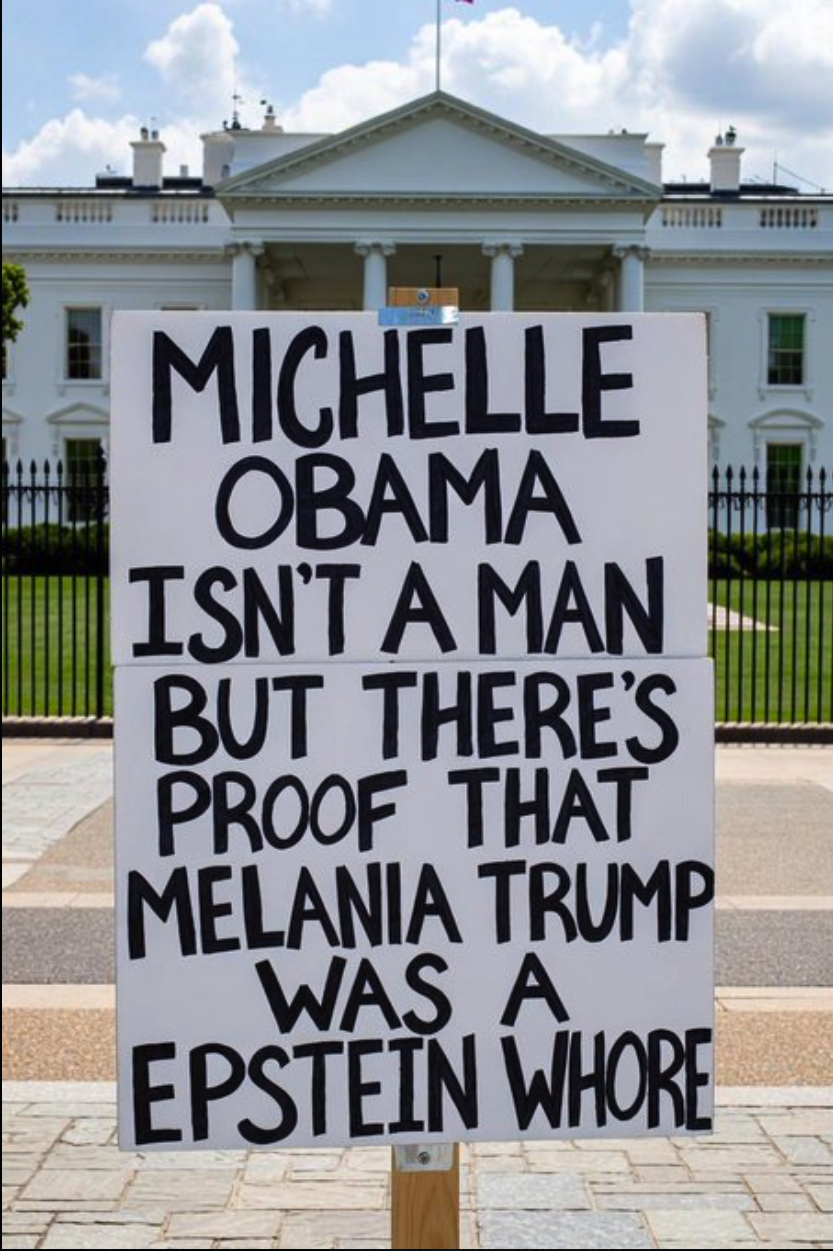 A handwritten protest sign mounted on a wooden post on the lawn in front of the White House.