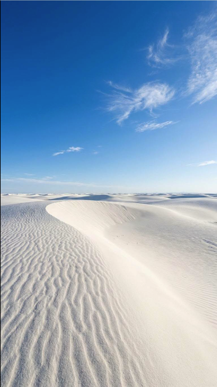 Rolling white sand dunes stretching toward the horizon beneath a blue sky with scattered wispy clouds.