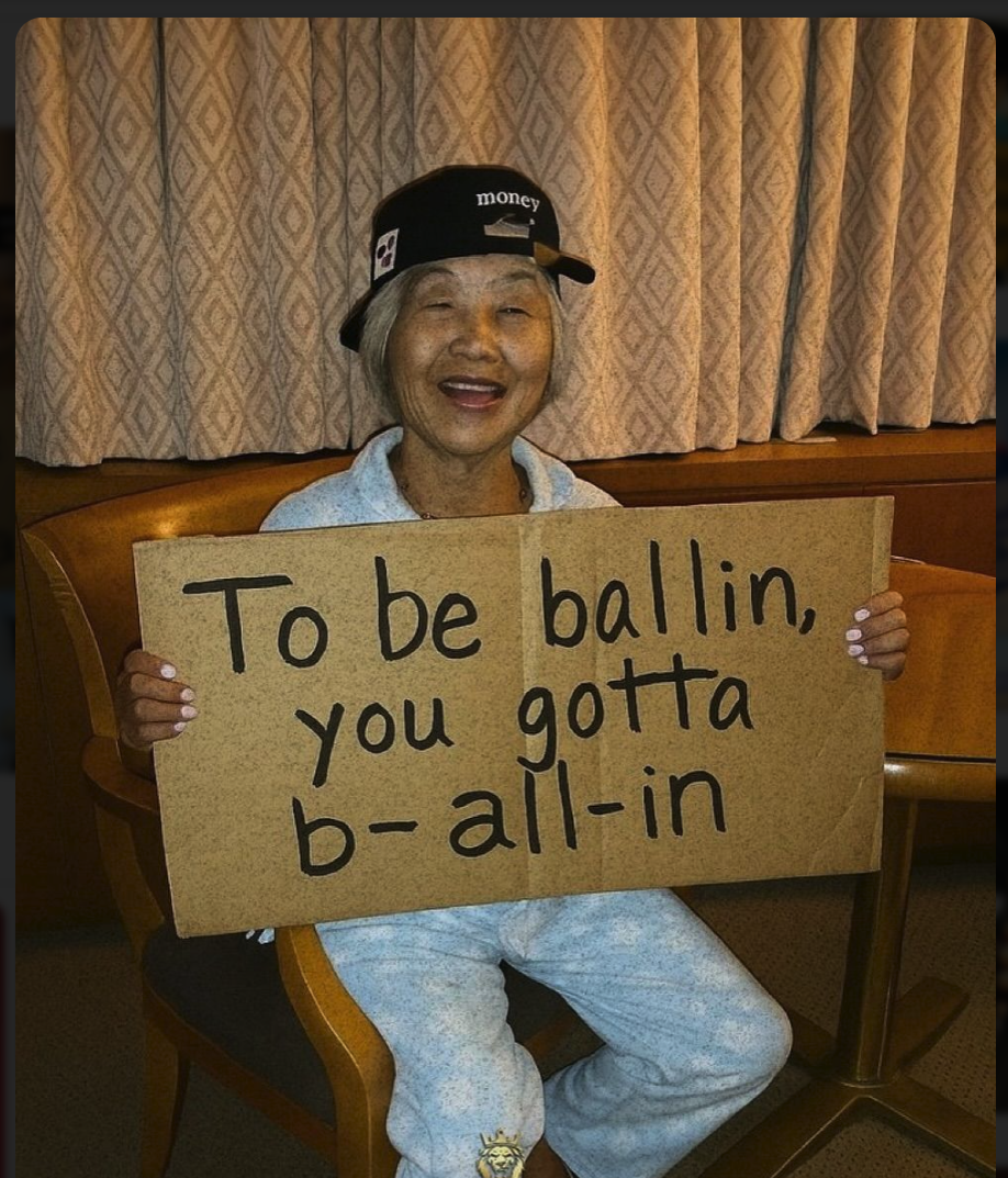 An elderly woman in loungewear and a cap reading 'money' holding a cardboard sign that says 'To be ballin, you gotta b- all-in'.