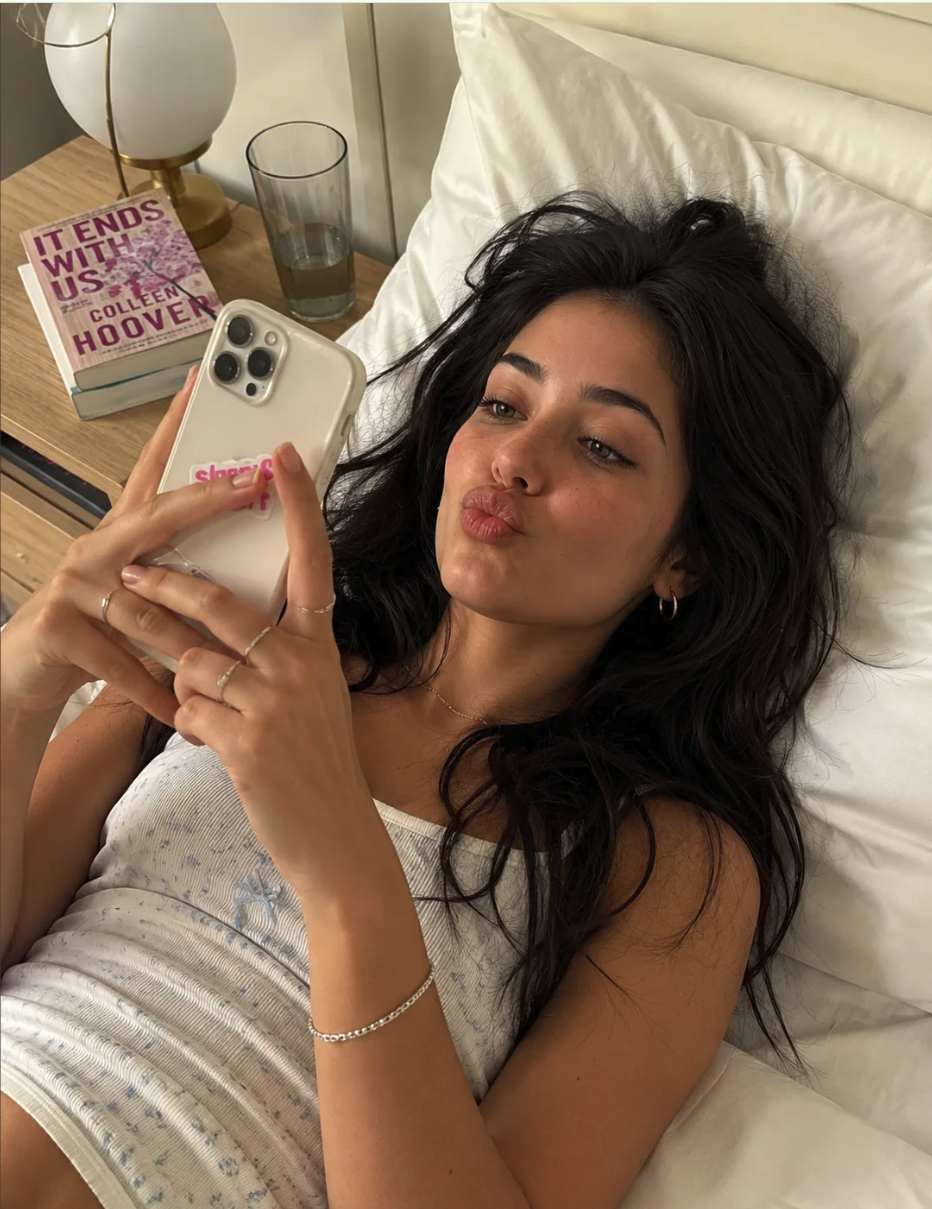 A woman with long dark hair lying in bed taking a selfie while holding a smartphone, with a copy of 'It Ends With Us' on the nightstand.