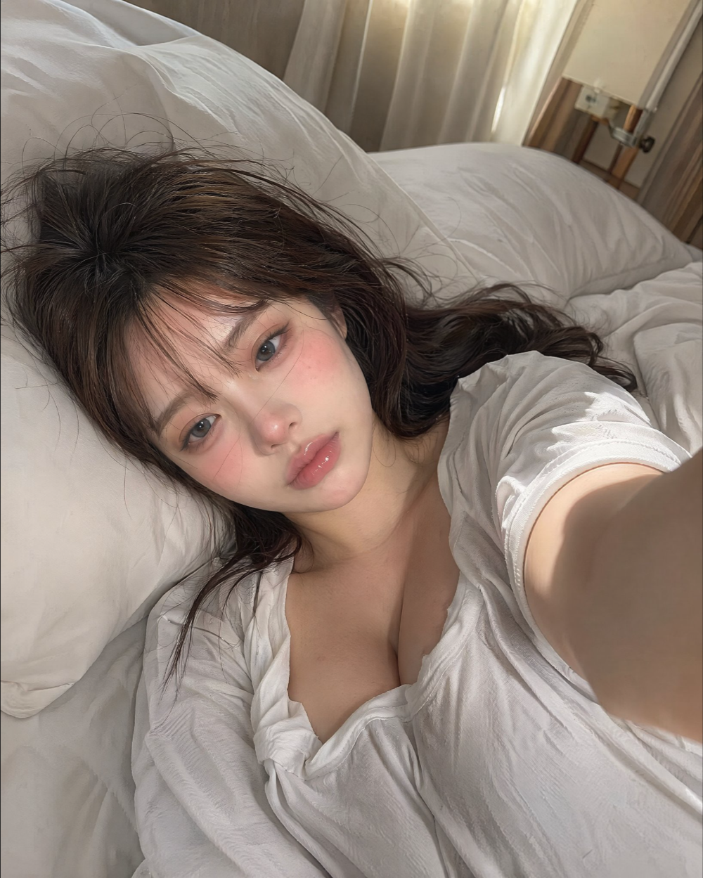 A young woman with dark hair lying on white bedding, taking a selfie in a loose white shirt.
