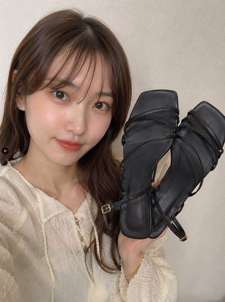 A woman with long brown hair holding up a pair of black strappy heeled sandals.