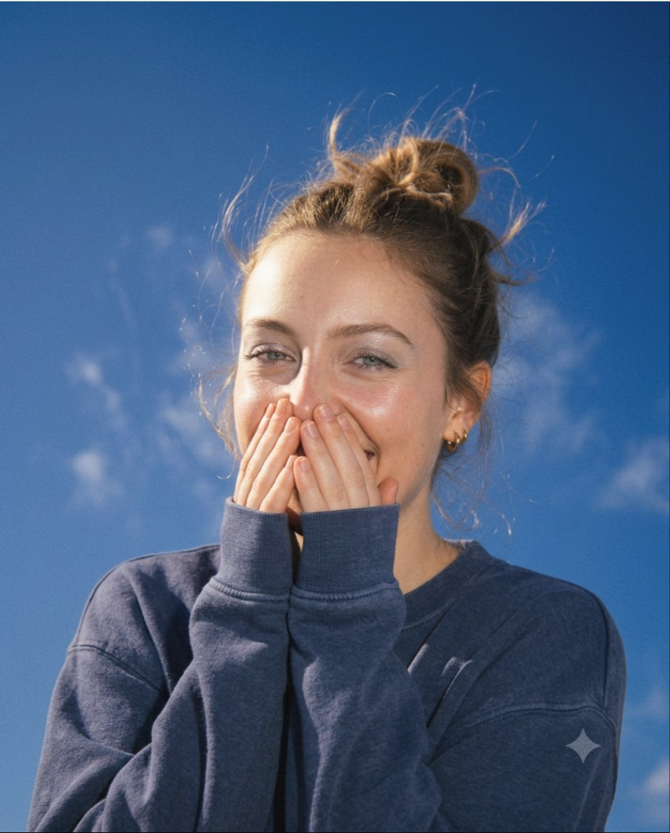 An unidentified young woman in a blue sweatshirt smiling with her hands cupped near her face against a bright blue sky.