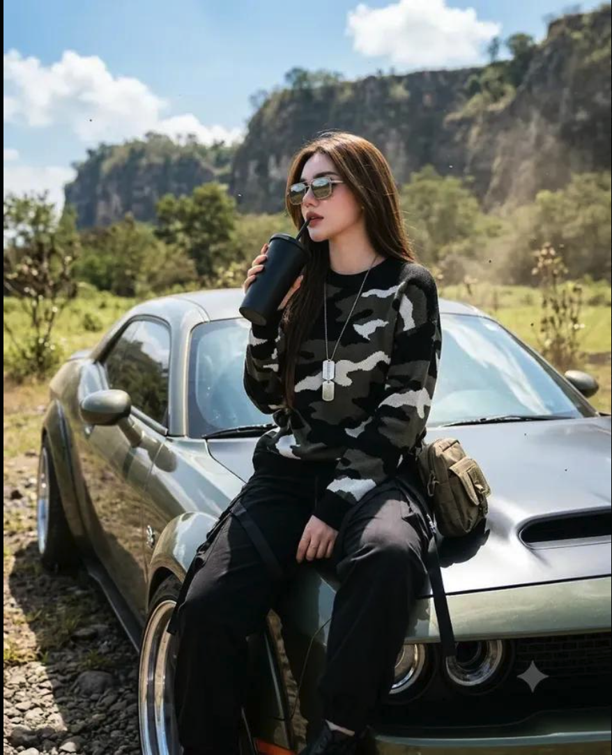 A woman with long dark hair, sunglasses, and a camouflage sweater sitting on the hood of an olive-green car and drinking from a black cup.