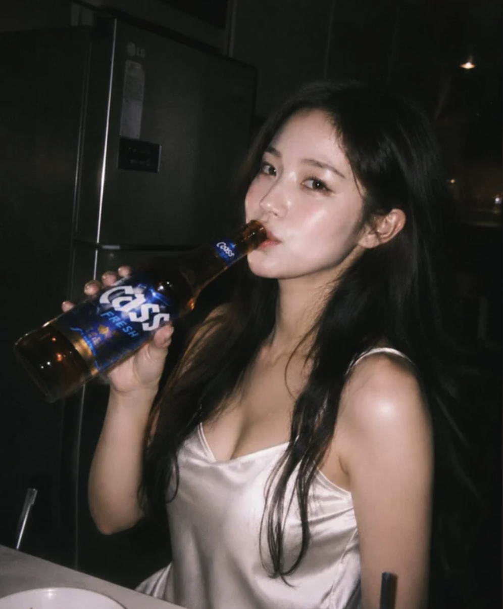 A young woman in a satin camisole drinking from a bottle of Cass Fresh beer in a dimly lit room.