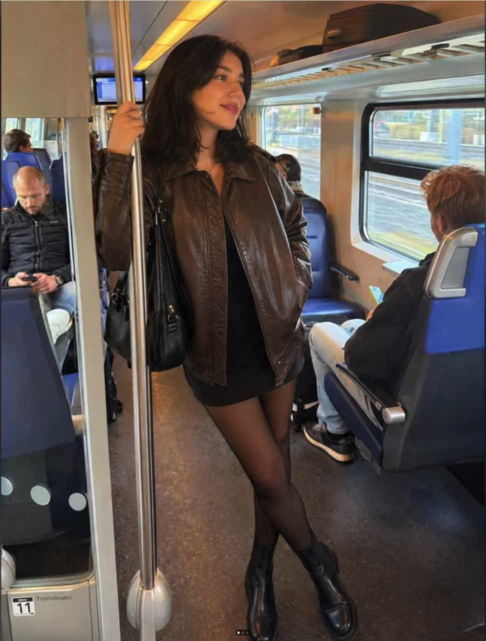 A woman in a brown leather jacket and tights standing on a train, holding a pole.