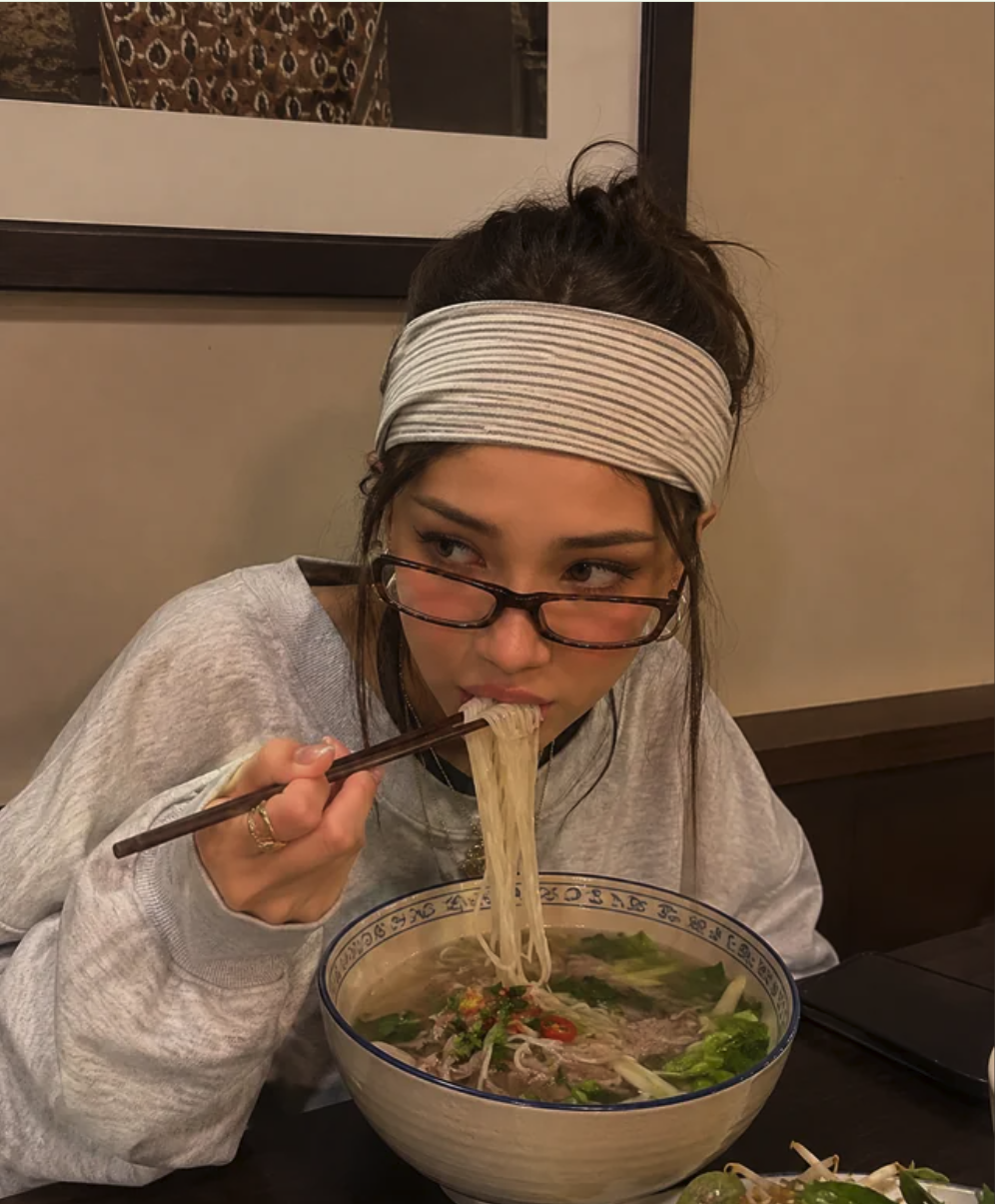 A woman wearing a striped headband and tinted glasses eating noodles from a large bowl with chopsticks at a restaurant table.
