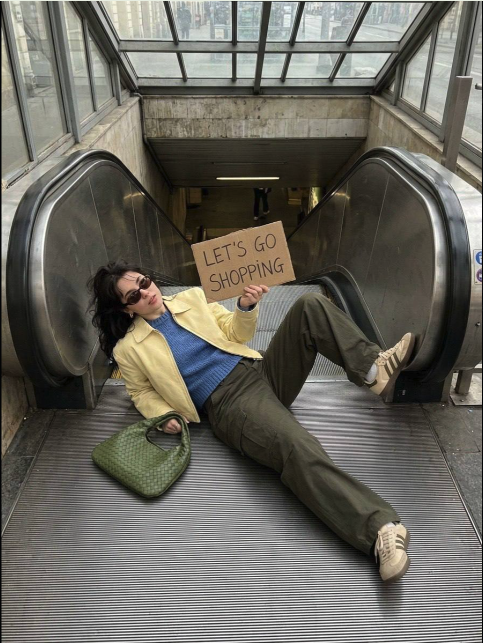 A woman in sunglasses reclining on a descending escalator, holding a cardboard 'Let's Go Shopping' sign.