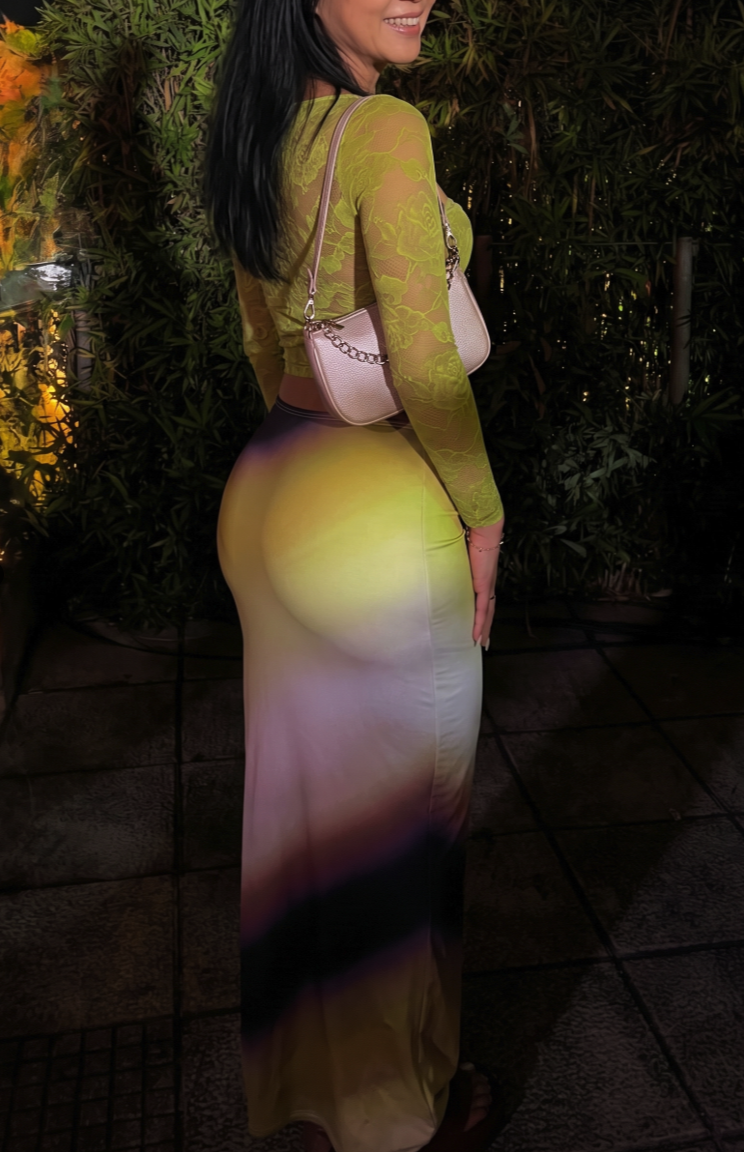 A woman with long dark hair wearing a green lace top and a long multicolored gradient skirt, photographed in profile outdoors at night.