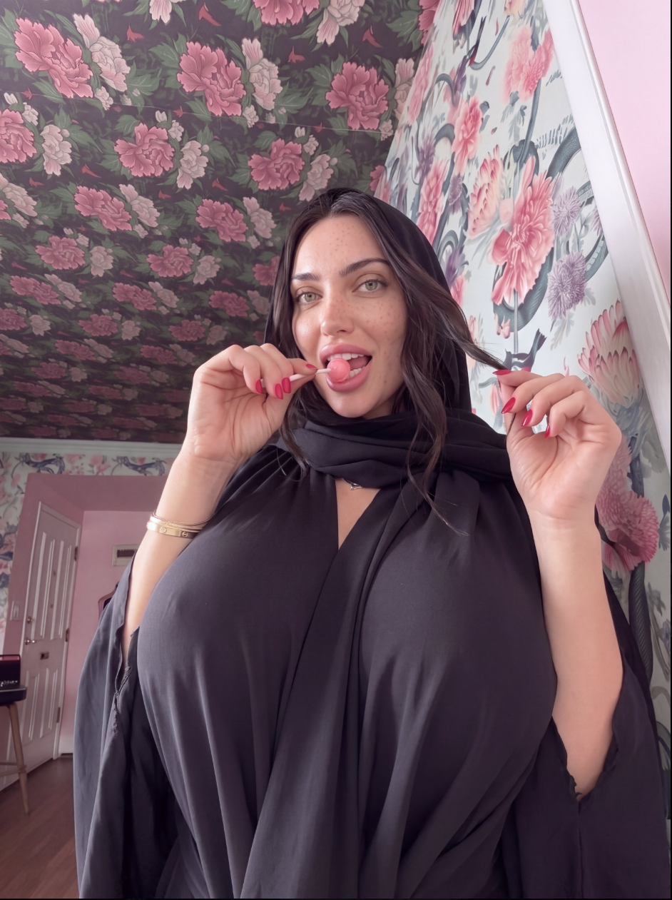 A woman in a black hijab and abaya holding a pink lollipop near her open mouth, with floral wallpaper behind her.