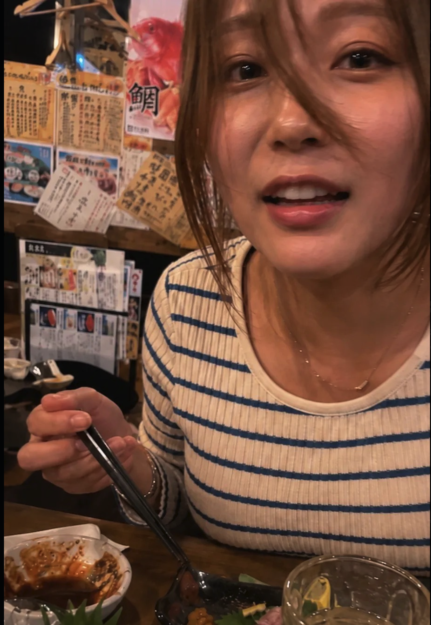 A woman with brown hair in a striped shirt, seated at a table holding chopsticks in a Japanese restaurant with menu posters on the wall behind her.