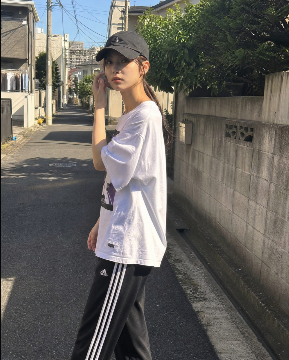 A woman in a black baseball cap, white t-shirt, and Adidas track pants standing on a narrow Japanese street, holding a phone to her ear.