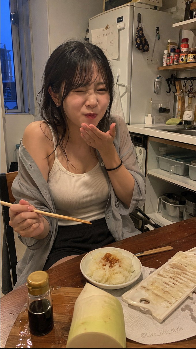 A young woman seated at a kitchen table blowing a kiss, with a bowl of rice and a white vegetable on the table in front of her.