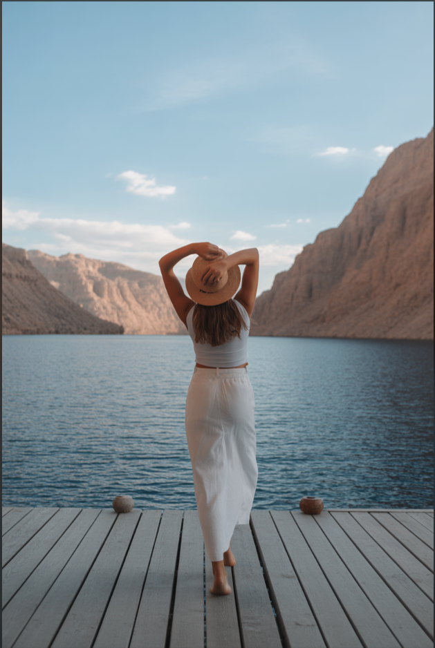 A woman in white clothing and a straw hat standing on a wooden deck facing a mountain lake.