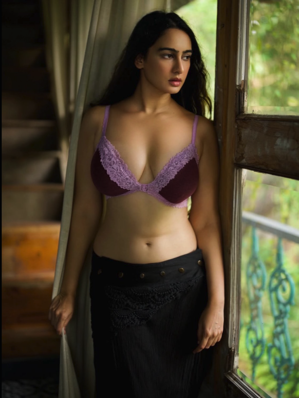 A woman in a purple lace bra and black skirt standing by a window.