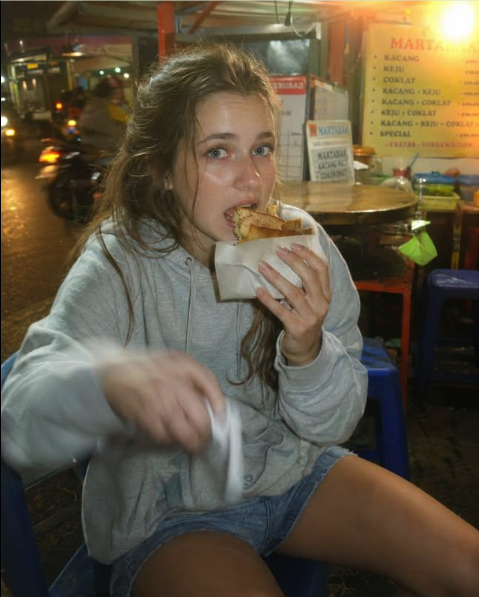 A woman in a gray hoodie appearing to eat a wrapped martabak at an Indonesian street food stall at night.