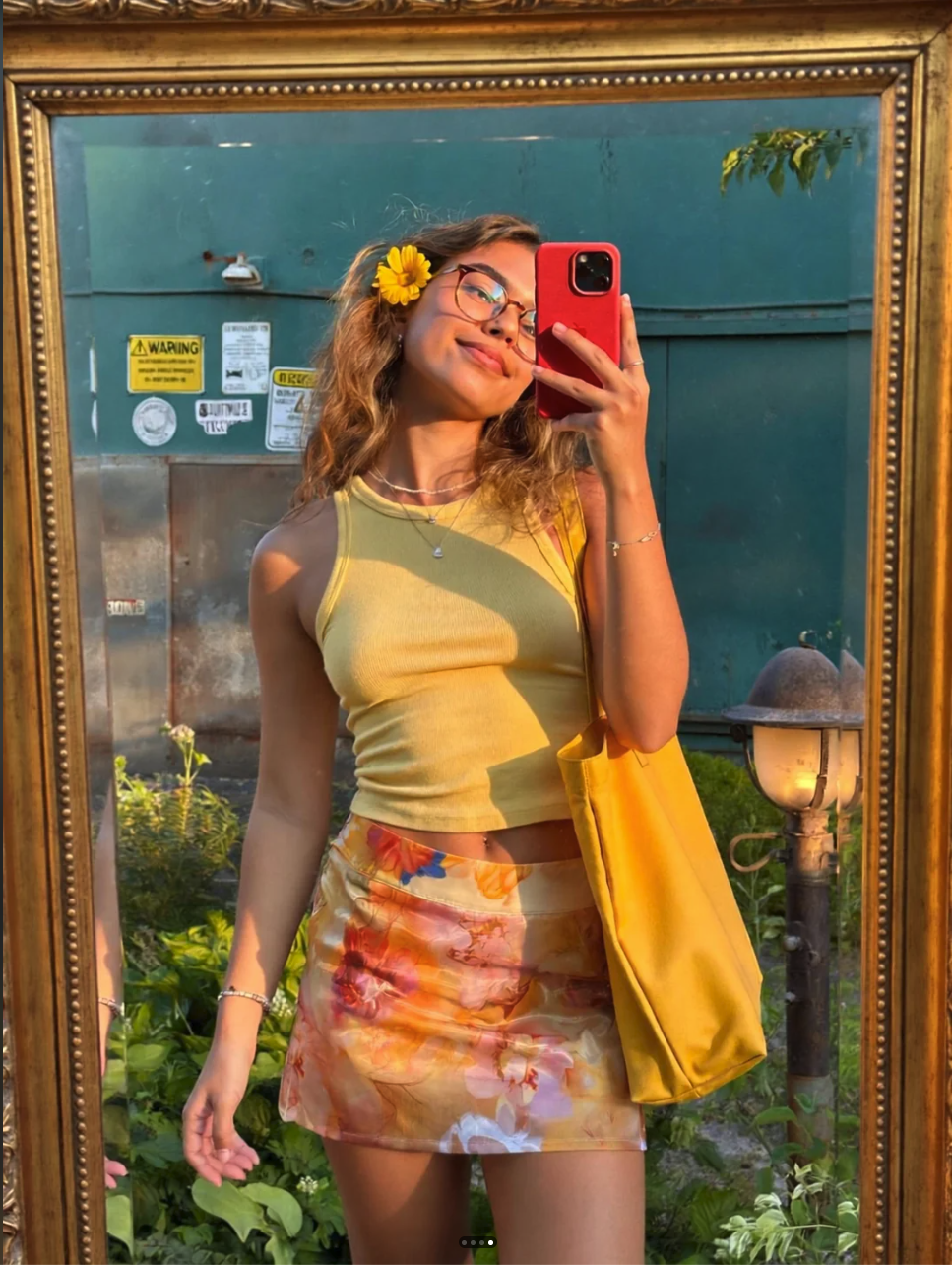 An unidentified woman in a yellow top and floral skirt taking a mirror selfie with a red smartphone.