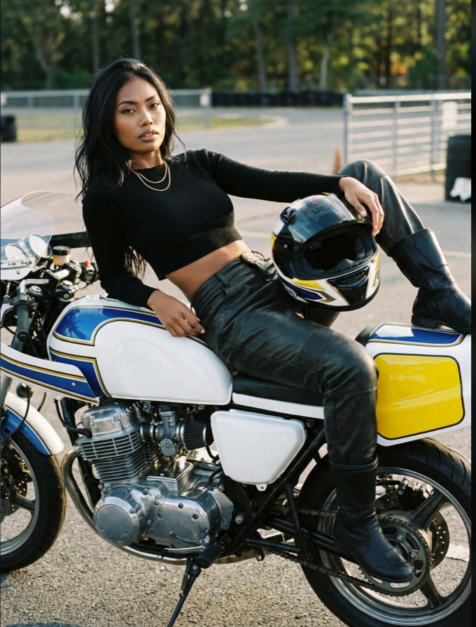 A woman with long dark hair in a black crop top and black leather pants reclining across a blue, white, and yellow motorcycle.