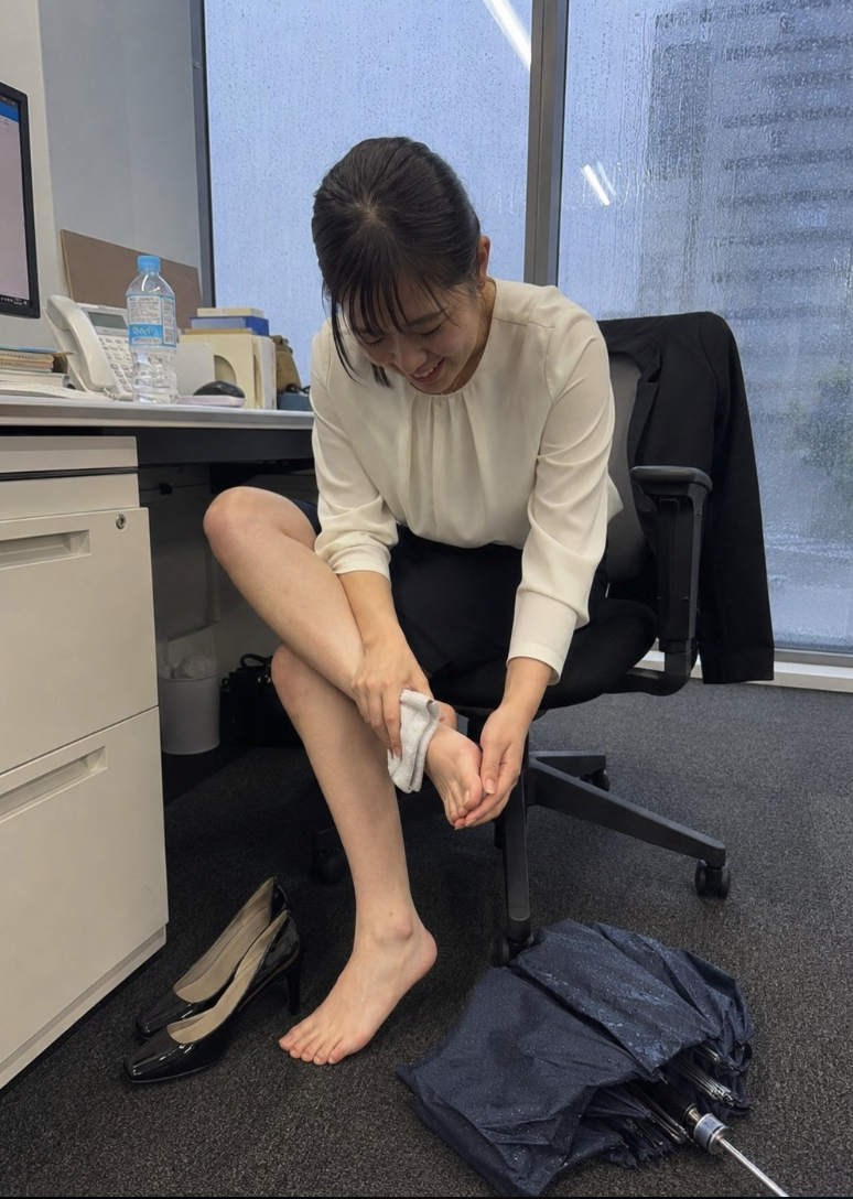 A woman in a cream blouse and dark skirt seated in an office chair, wiping her bare foot with a small cloth.