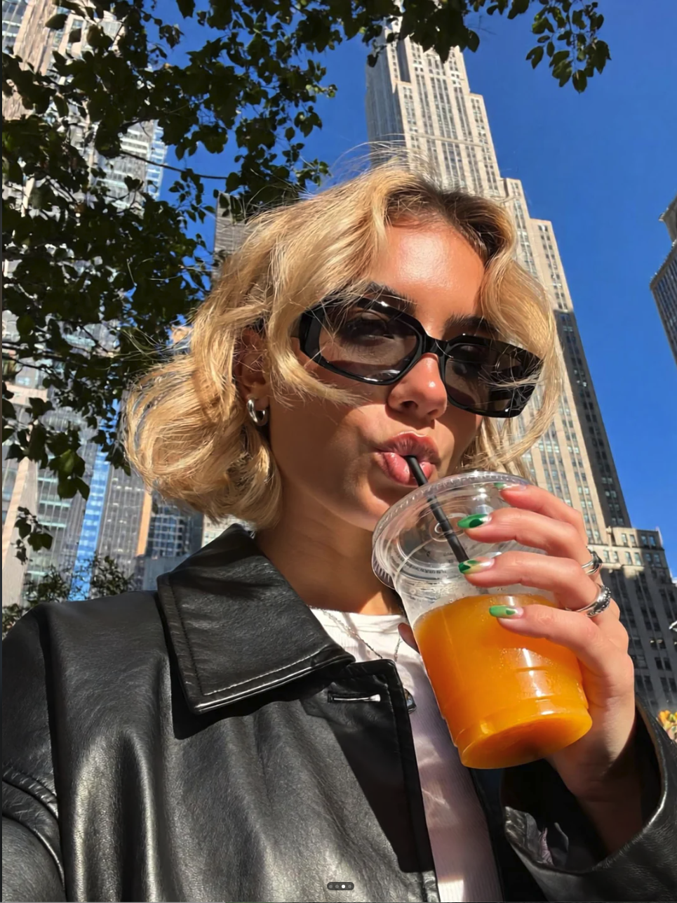 A woman with blonde hair and dark sunglasses drinking from a clear plastic cup against an urban skyscraper backdrop.