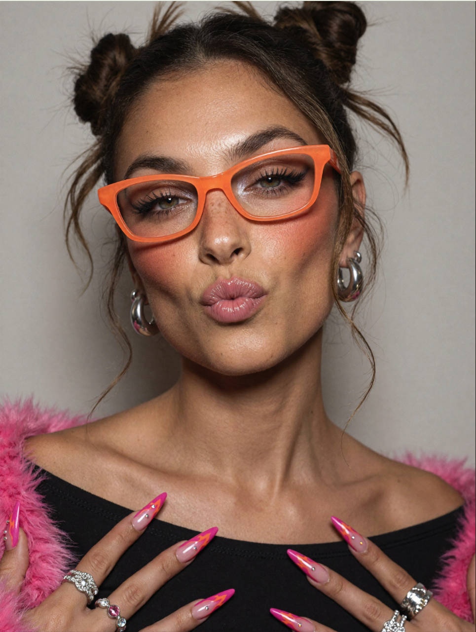 A close-up portrait of a woman wearing orange cat-eye glasses and a pink fluffy garment, puckering her lips.