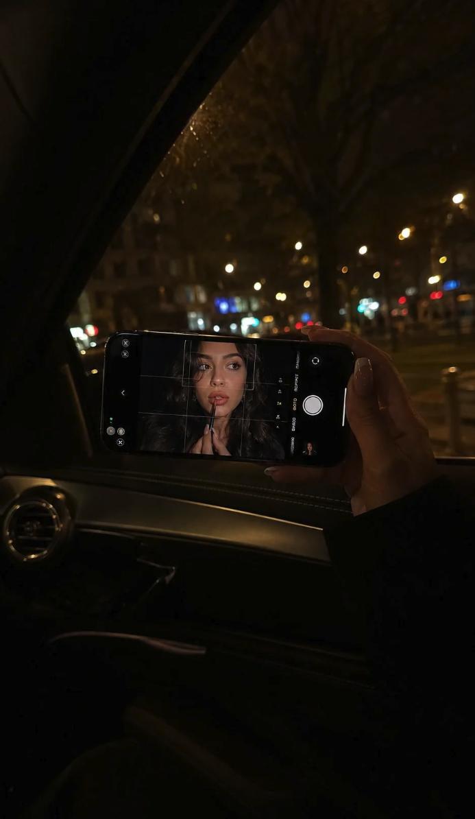A smartphone held at night displaying a camera viewfinder with a woman applying lip gloss visible on the screen.